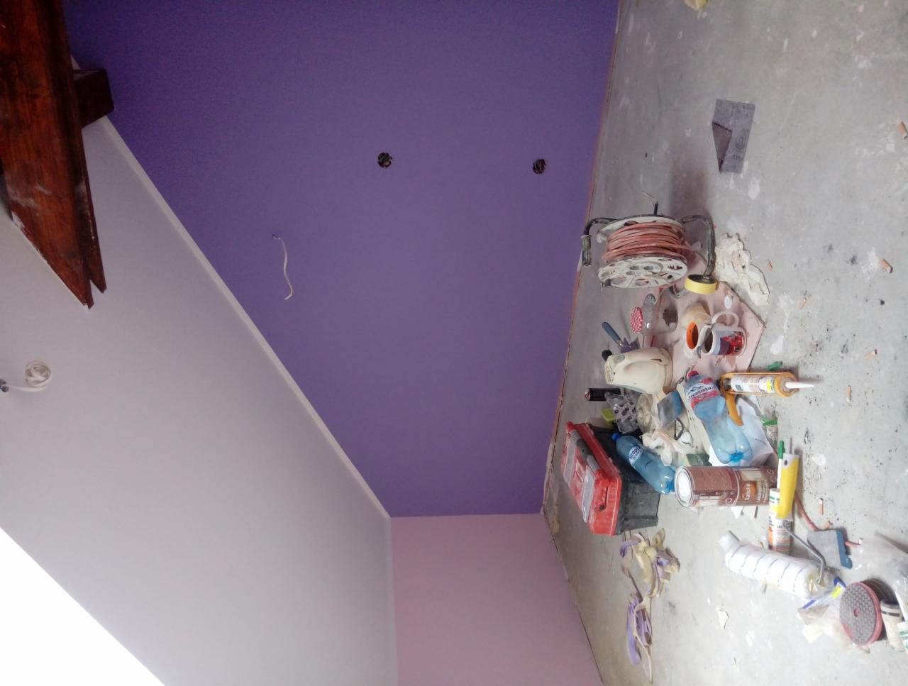 Fioletowy sufit świeżo po malowaniu, widoczne narzędzia malarskie i materiały budowlane na podłodze, surowy stan wykończenia.