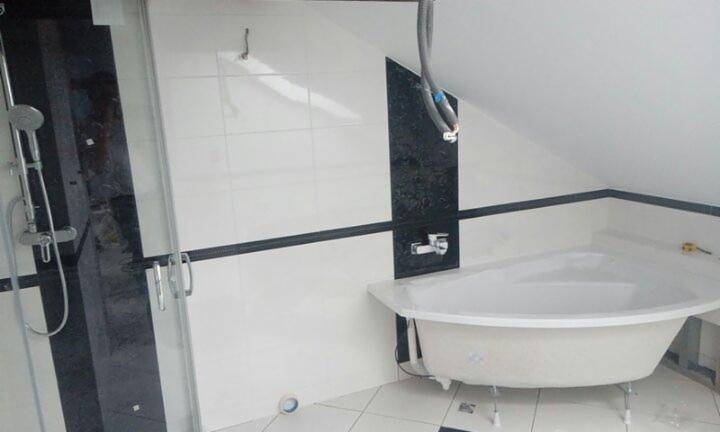 Narożna wanna na nóżkach w łazience z biało-czarnymi płytkami, widoczna kabina prysznicowa z chromowaną armaturą i fragment skośnego sufitu.