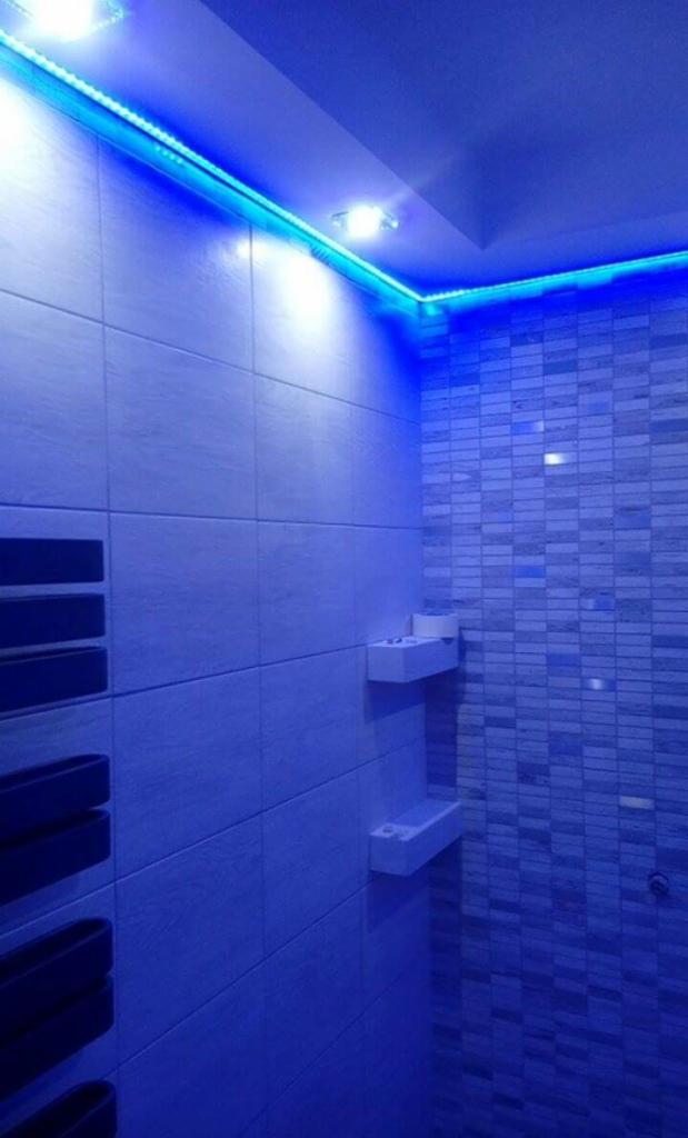 Nowoczesna łazienka z niebieskim oświetleniem LED, białe półki na ścianie, płytki imitujące cegłę na ścianie prysznicowej.