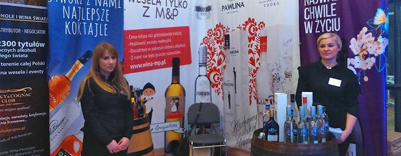 Stoisko promocyjne z alkoholem na targach, z bannerami reklamowymi, prezentacją butelek na beczce i dwiema kobietami obsługującymi stoisko.