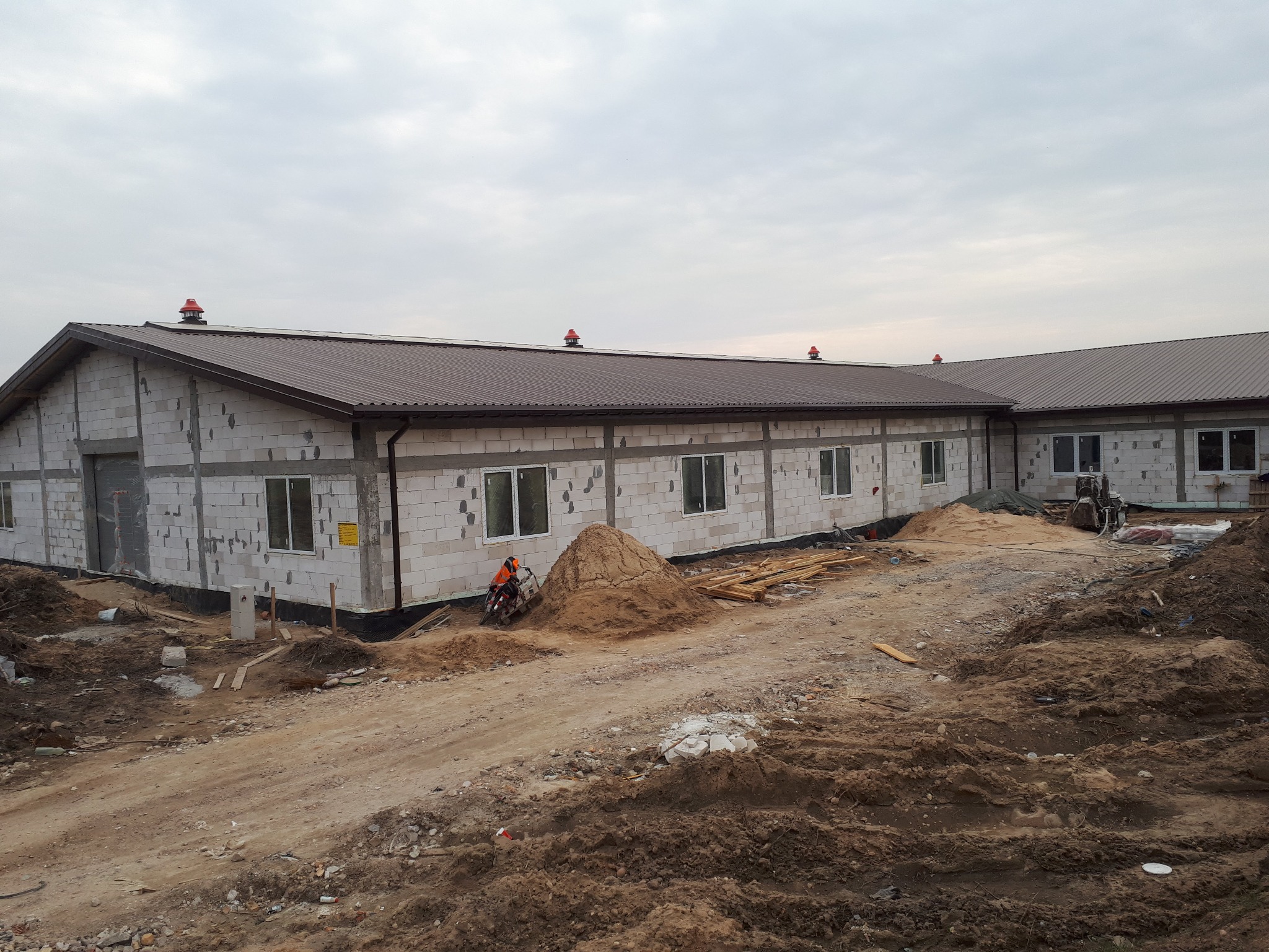 Budynek w trakcie budowy z widoczną konstrukcją dachu pokrytą blachodachówką w kolorze brązowym, z czerwonymi wywietrznikami kominowymi, ściany z białych bloczków, teren wokół budynku częściowo...