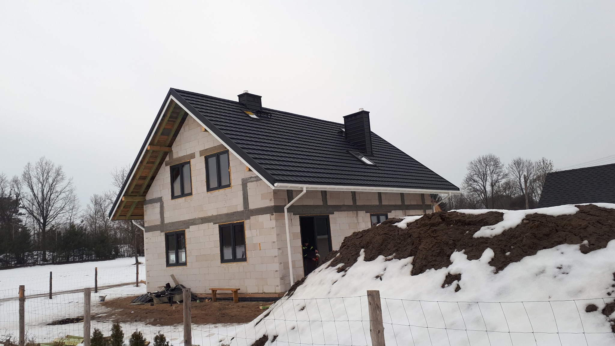 Nowy dom w trakcie budowy z czarnym dachem z blachy, widoczne kominy, orynnowanie, ściany z białego bloczka i częściowo ośnieżona sterta ziemi przed budynkiem.