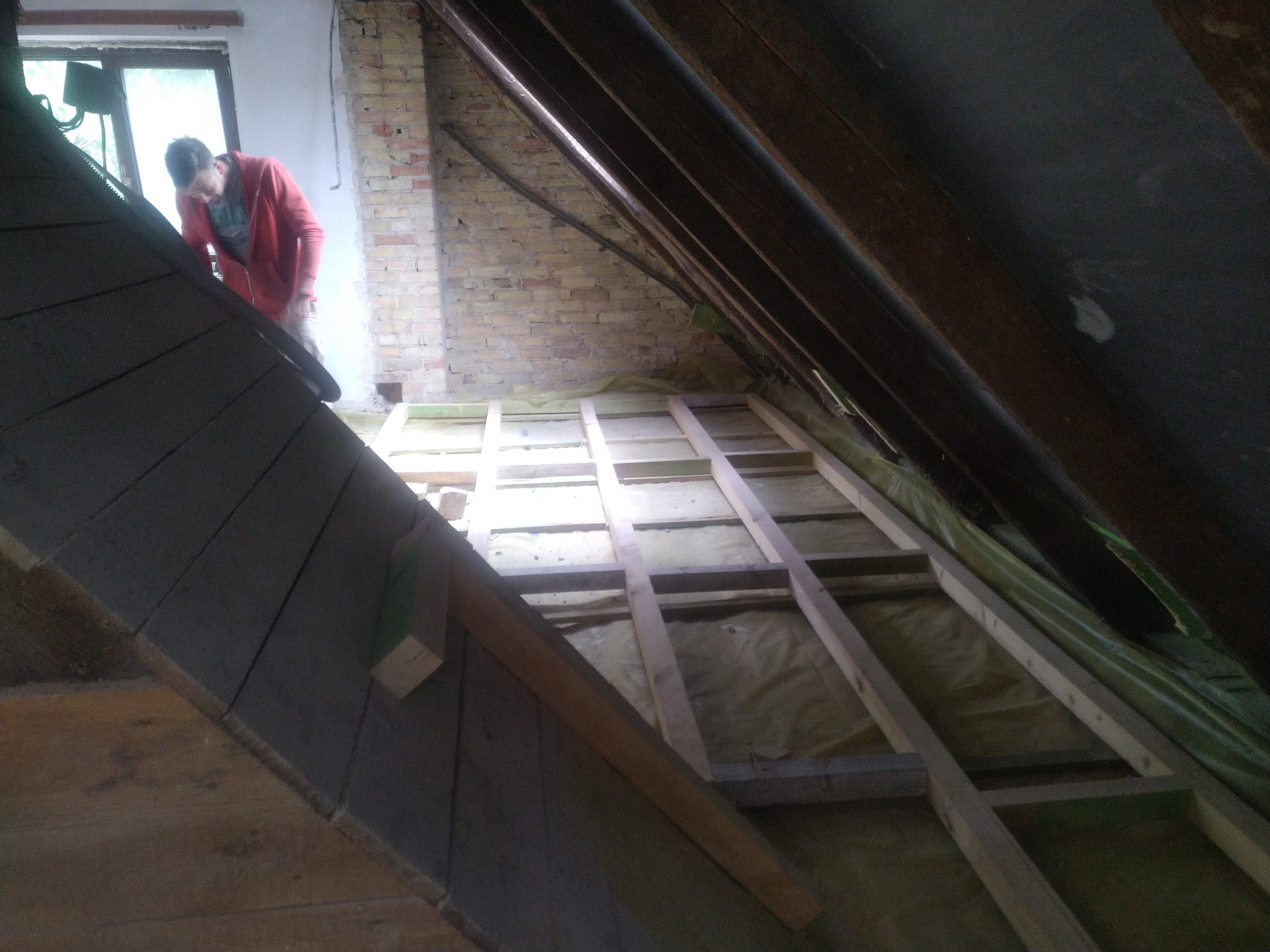 Montaż izolacji poddasza: widok na drewnianą konstrukcję szkieletową, wypełnioną materiałem izolacyjnym, z pracownikiem w czerwonej bluzie w tle przy oknie.