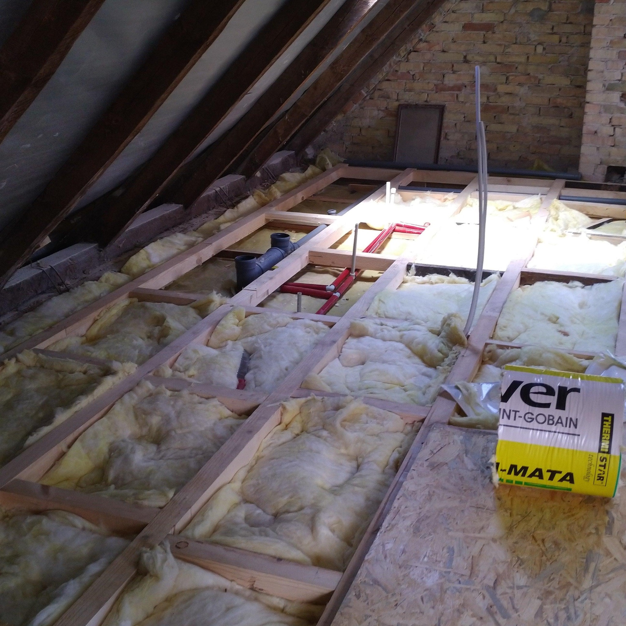 Poddasze w trakcie izolacji termicznej: wełna mineralna między drewnianymi belkami, widoczne rury instalacji, karton z wełną Saint-Gobain na podłodze z płyt OSB, fragment muru z cegły w tle.