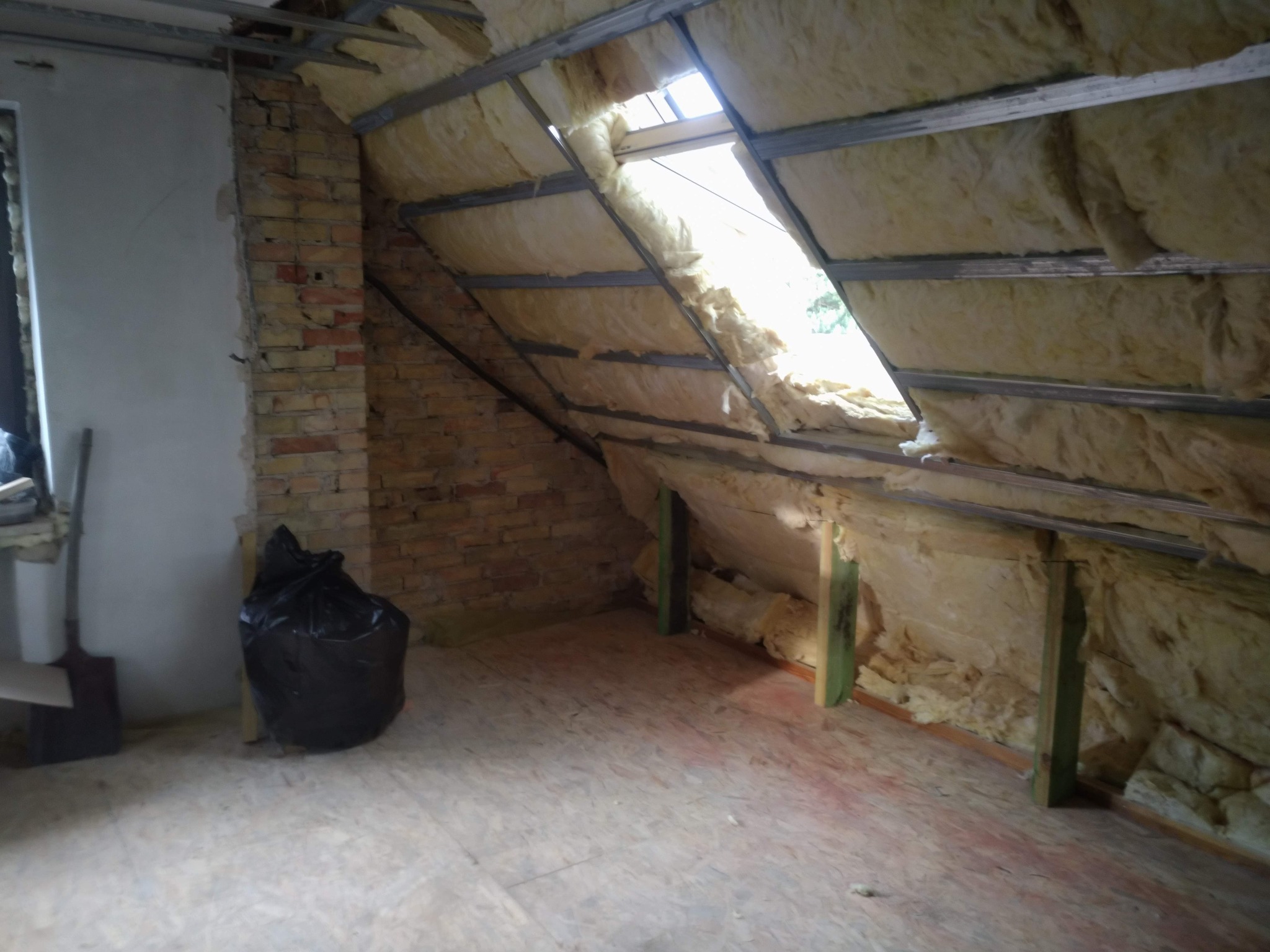 Poddasze w trakcie prac ociepleniowych, widoczne metalowe profile, wełna mineralna, fragment okna dachowego, ceglana ściana i czarny worek na śmieci.