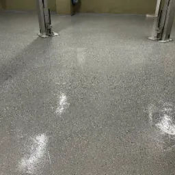 Błyszcząca, szara posadzka z drobnymi, ciemnymi plamkami w przestronnym garażu z podnośnikami samochodowymi. Gładka powierzchnia, widoczne odbicia światła.