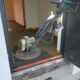 MP SYSTEM Piotr Marciniak - Maszyna do wygładzania posadzek jastrychowych w progu drzwi. Widoczna świeżo wylana, gładka posadzka z żółtą taśmą przy ścianach. Kabel zasilający podłączony do listwy.