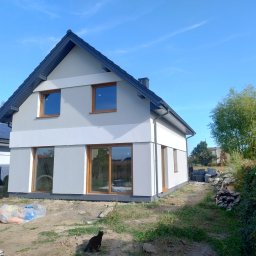 MP SYSTEM Piotr Marciniak - Nowoczesny dom jednorodzinny z szarą elewacją i drewnianymi oknami w słoneczny dzień. Na pierwszym planie czarny kot i materiały budowlane.