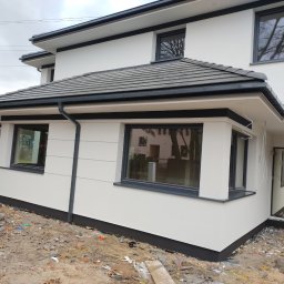 MP SYSTEM Piotr Marciniak - Nowoczesny dom z białą elewacją i grafitowymi detalami, widoczny z boku. Uwagę zwraca staranne wykończenie i kontrast kolorów. Teren wokół domu w trakcie prac wykończeniowych.