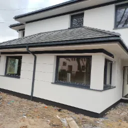 Nowoczesny dom z białą elewacją i grafitowymi detalami, widoczny z boku. Uwagę zwraca staranne wykończenie i kontrast kolorów. Teren wokół domu w trakcie prac wykończeniowych.