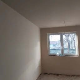 Wnętrze pokoju po tynkowaniu maszynowym, widoczne gładkie ściany w neutralnym odcieniu, okno z widokiem na zewnątrz i surowa podłoga przed wylewką.