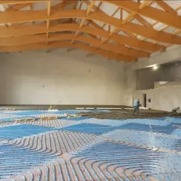 Wnętrze hali sportowej z ułożonym systemem ogrzewania podłogowego w postaci niebieskich rurek na siatce, częściowo pokrytych mokrą wylewką, drewniana konstrukcja dachu