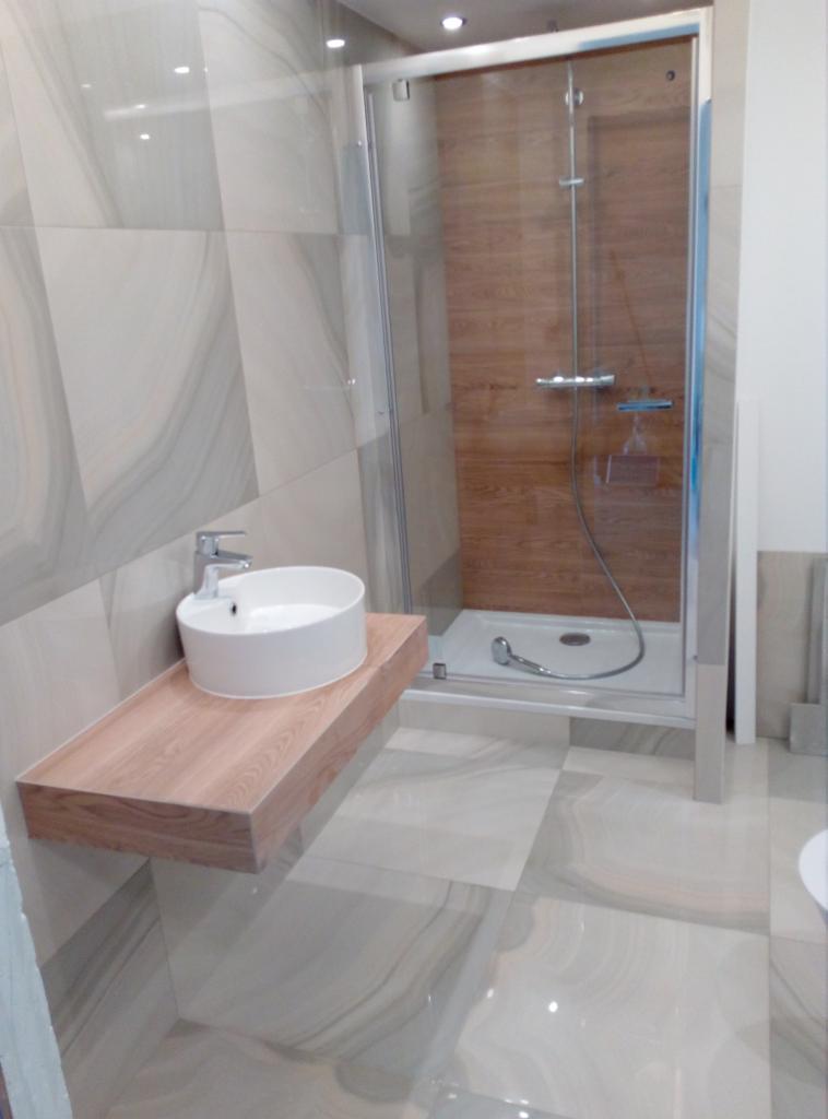 Nowoczesna łazienka z kabiną prysznicową z drewnianym panelem i okrągłą umywalką nablatową na drewnianym blacie, płytki podłogowe i ścienne imitujące marmur.