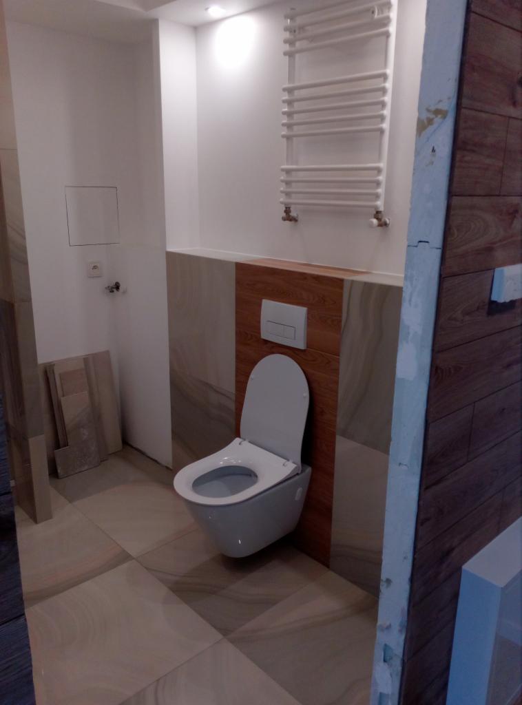 Zamontowana podwieszana toaleta z deską w łazience z płytkami imitującymi drewno, widoczny grzejnik drabinkowy i fragment ściany w trakcie wykończenia.