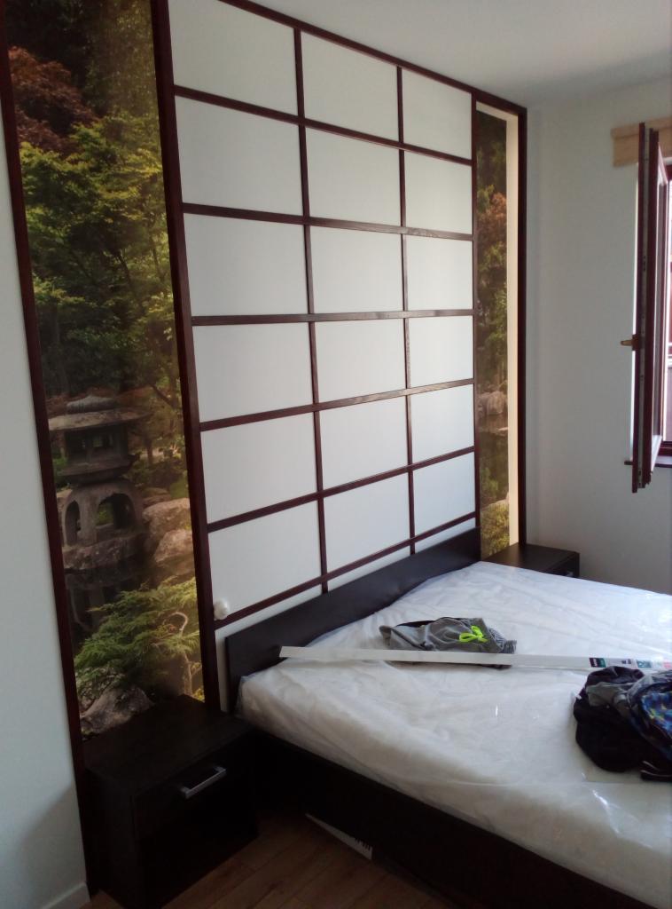 Sypialnia z łóżkiem i panelem ściennym w stylu japońskim z motywem ogrodu i pagody, widok na szafkę nocną i okno.