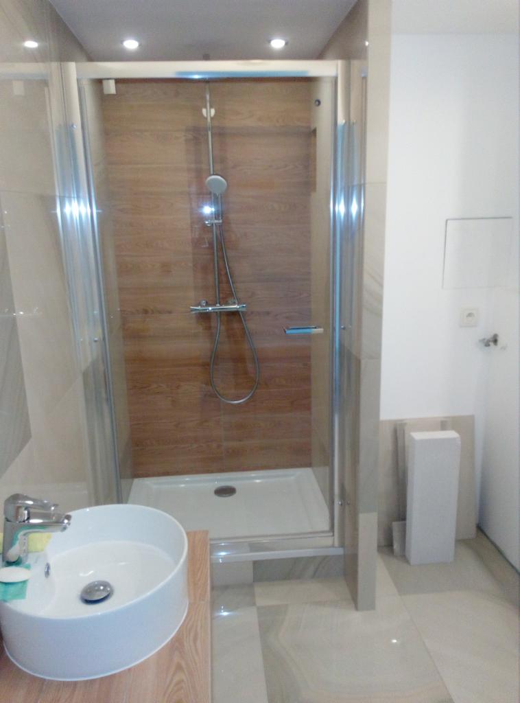 Nowoczesna kabina prysznicowa z panelami imitującymi drewno, okrągła umywalka z chromowaną baterią, jasne płytki podłogowe i białe ściany w łazience po remoncie.