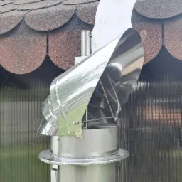 Błyszczący, metalowy wiatrochron kominowy z motywem dekoracyjnym na szczycie, zamontowany na kominie z brązowym dachem w tle.