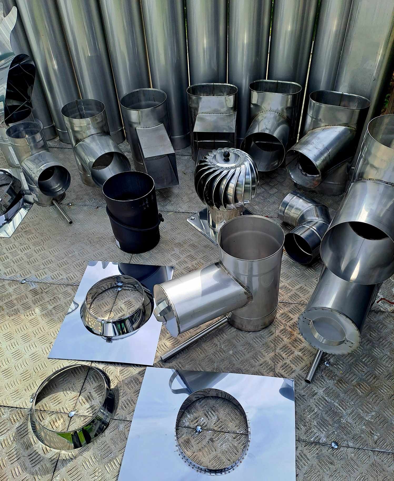 Różne elementy ze stali nierdzewnej do systemów odprowadzania spalin, w tym rury, kolanka, rozety i nasada turbinowa, ułożone na metalowej powierzchni o wzorze rombów.