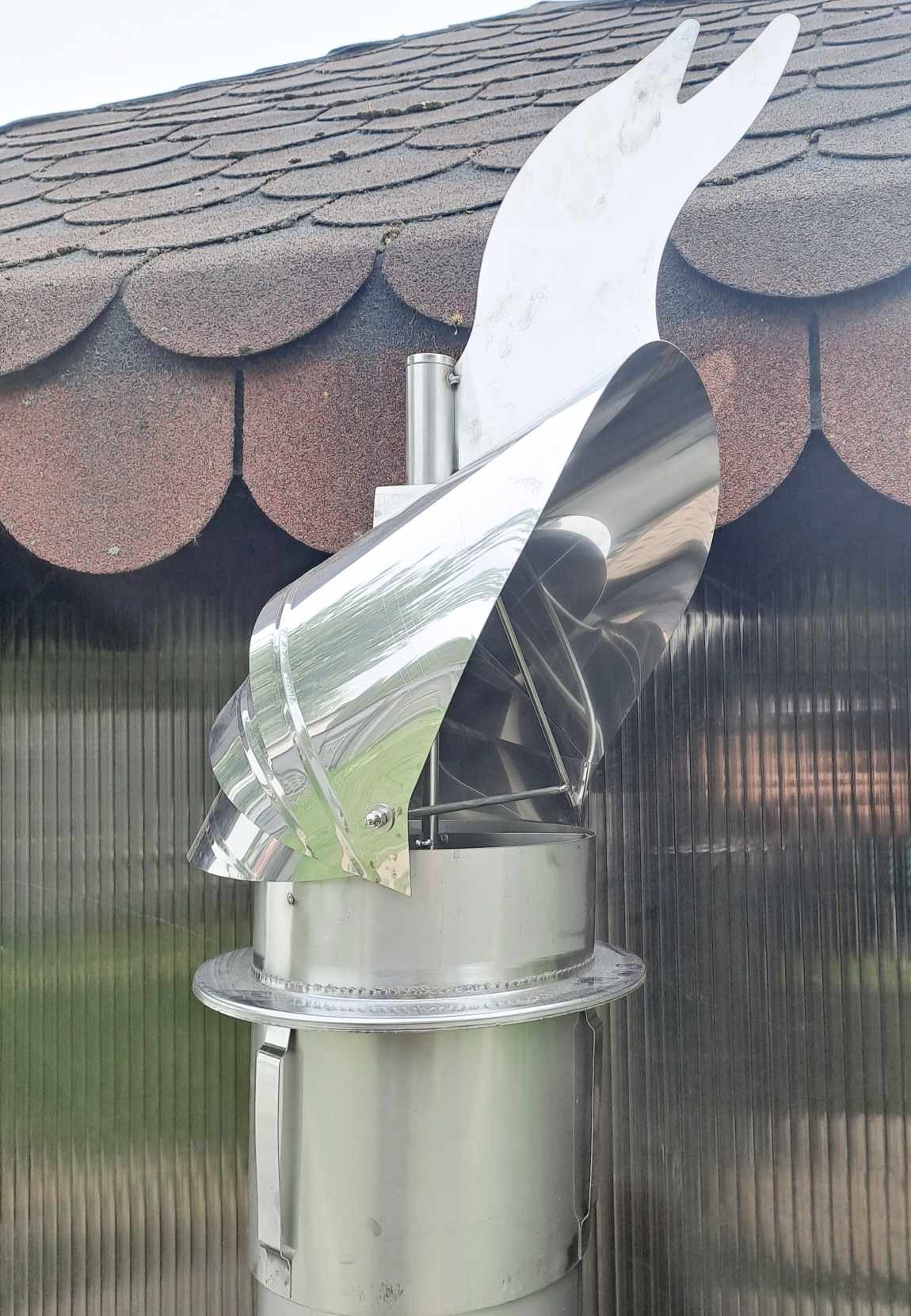 Błyszczący, metalowy wiatrochron kominowy z motywem dekoracyjnym na szczycie, zamontowany na kominie z brązowym dachem w tle.