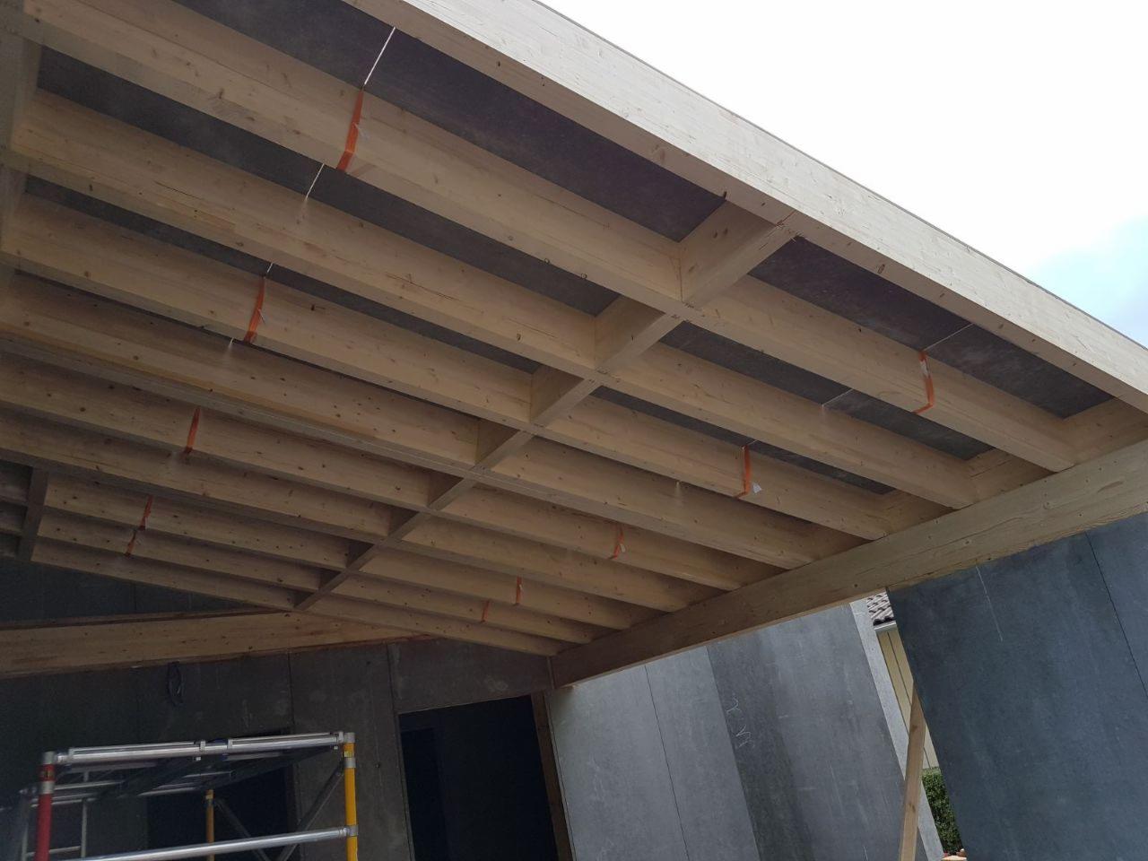 Konstrukcja stropu z belek drewnianych i płyt OSB w trakcie montażu, widoczne pomarańczowe taśmy mocujące, szare ściany budynku w surowym stanie.