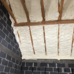 Poddasze ocieplone pianką poliuretanową, widoczne drewniane belki konstrukcyjne i fragment ściany z szarego bloczka betonowego.