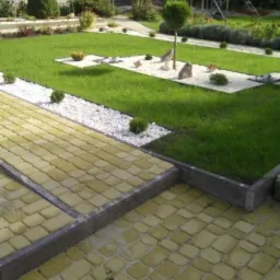 Uporządkowany ogród z żółtą kostką brukową, trawnikiem, białymi kamieniami i niskimi krzewami, widok z góry.