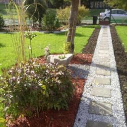 Justyna Denis-Makara - Ścieżka z płyt betonowych i białego grysu w ogrodzie, obrzeża z brązowej kory, w tle trawnik i drzewa.