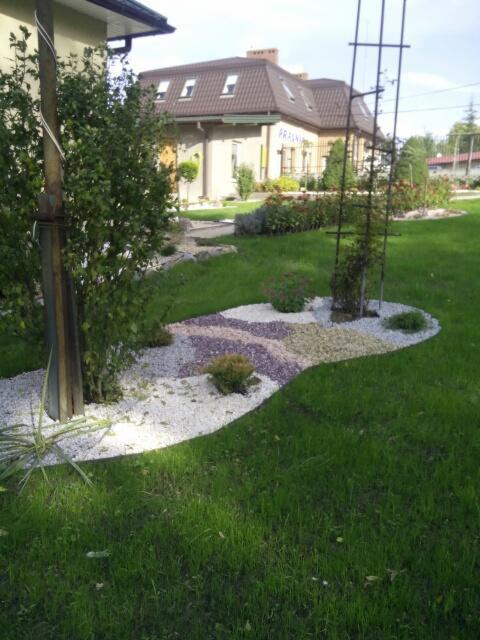 Ogród z ozdobnymi rabatami wysypanymi białym, fioletowym i beżowym żwirem, widoczna trawa i budynek w tle.