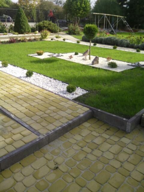 Uporządkowany ogród z żółtą kostką brukową, trawnikiem, białymi kamieniami i niskimi krzewami, widok z góry.