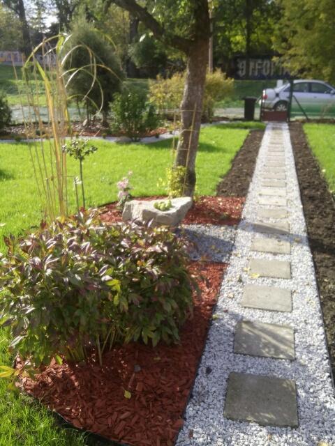 Ścieżka z płyt betonowych i białego grysu w ogrodzie, obrzeża z brązowej kory, w tle trawnik i drzewa.