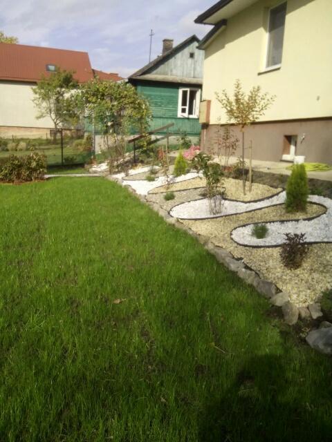 Ujęcie ogrodu z świeżo posianą trawą, ozdobnymi rabatami z białego i brązowego żwiru, młodymi drzewkami i krzewami, obrzeżami z kamieni polnych, na tle zabudowy mieszkalnej.