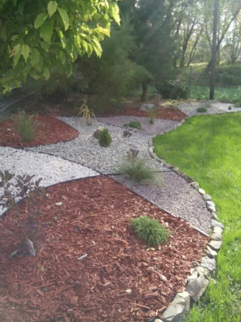 Rabata z kory i jasnych kamieni oddzielona kamiennym obrzeżem, obok trawnik i drzewa w tle.