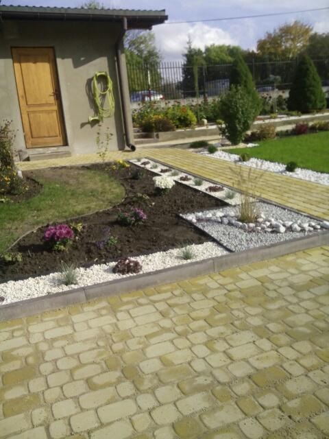 Uporządkowany ogród z nowo posadzonymi kwiatami i dekoracyjnymi kamieniami, widok na kostkę brukową i budynek gospodarczy z wężem ogrodowym.