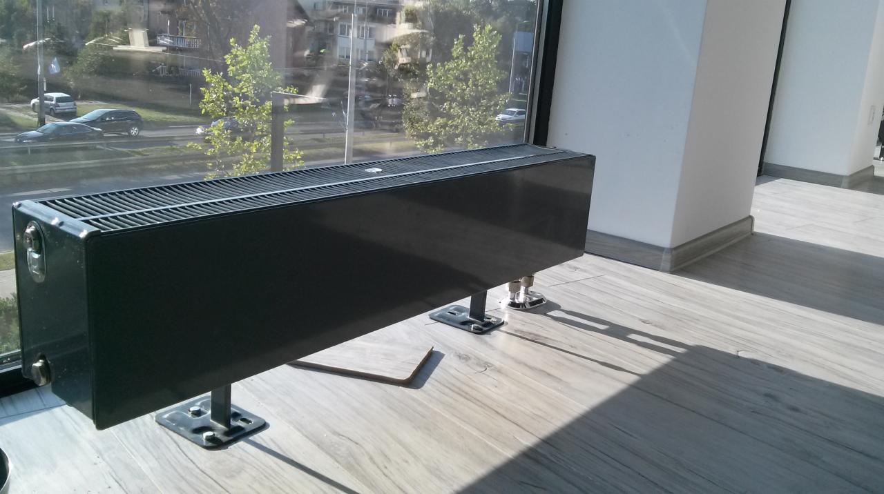 Czarny, niski grzejnik kanałowy zamontowany przy oknie z widokiem na ulicę, stojący na drewnopodobnej podłodze.