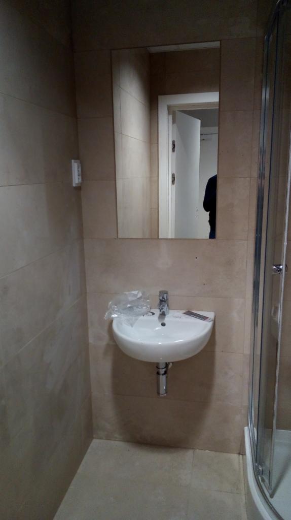 Nowa, biała umywalka zamontowana w łazience z beżowymi płytkami, zabezpieczona folią ochronną, widoczne rury odpływowe i fragment kabiny prysznicowej z przezroczystego szkła.