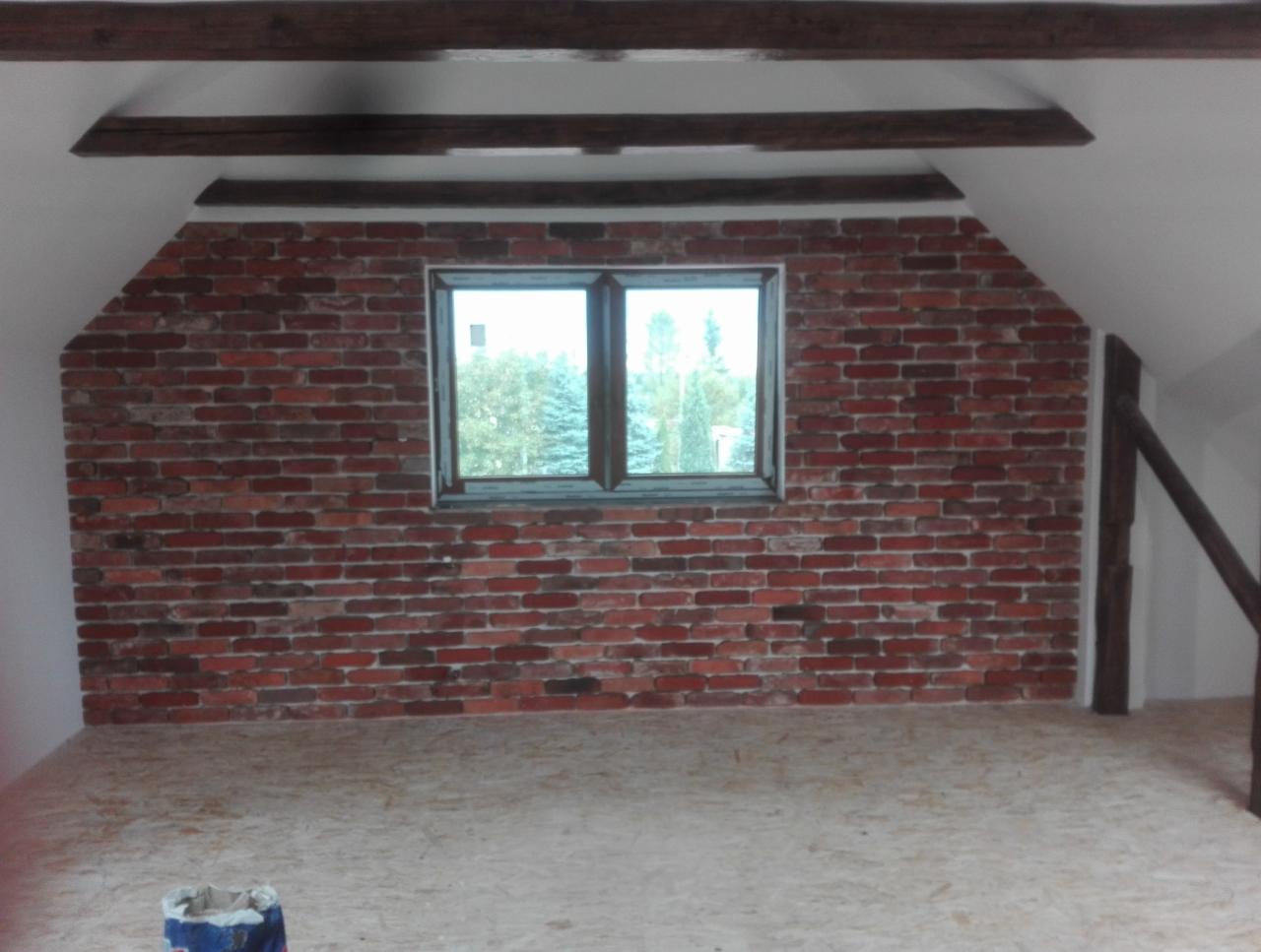 Poddasze z oknem, ściana wykończona czerwoną cegłą, drewniane belki stropowe, podłoga z płyt OSB, widoczny fragment balustrady schodów
