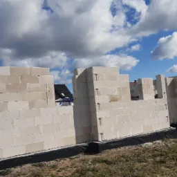 Wznoszenie ścian domu z białych bloczków betonowych, widoczne zbrojenie pionowe, czarna izolacja fundamentów, w tle niebo z chmurami i fragment dachu sąsiedniego budynku.