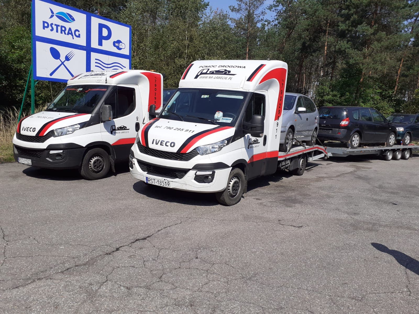 Dwie białe lawety Iveco z czerwono-czarnymi akcentami, jedna z dwoma samochodami na platformie, zaparkowane na parkingu przy lesie i znaku 'Pstrąg P WiFi'.
