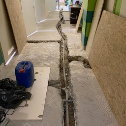 Specdom - Korytarz w trakcie prac instalacyjnych: wykopane bruzdy w podłodze, widoczne rury, narzędzia i materiały budowlane. W tle osoba wchodząca do pomieszczenia.