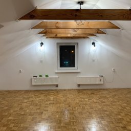 Specdom - Poddasze po remoncie: parkiet w jodełkę, białe ściany, drewniane belki stropowe z lampami, okno. Widoczne grzejniki i gniazdka elektryczne. Noc za oknem.