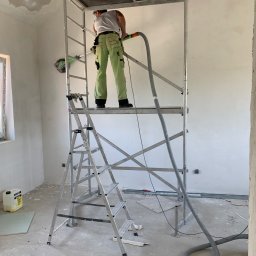 Specdom - Pracownik na rusztowaniu szlifuje ścianę w jasnym wnętrzu. Widoczny odkurzacz przemysłowy i drabina. Ściany przygotowane do malowania. Etap prac wykończeniowych.