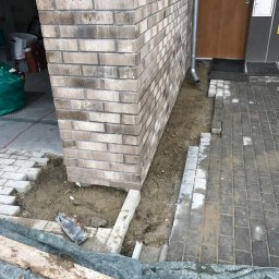 Specdom - Układanie kostki brukowej przy ścianie budynku z cegły. Widoczne fragmenty ułożonej kostki, piasek i narzędzia. Prace brukarskie w toku, z bliska.