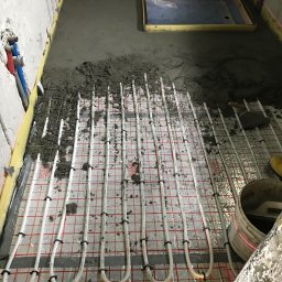 Specdom - Montaż ogrzewania podłogowego w łazience: białe rurki na folii, częściowo zalane betonem, widoczny laser krzyżowy i wiadro z zaprawą. Etap prac przed położeniem płytek.