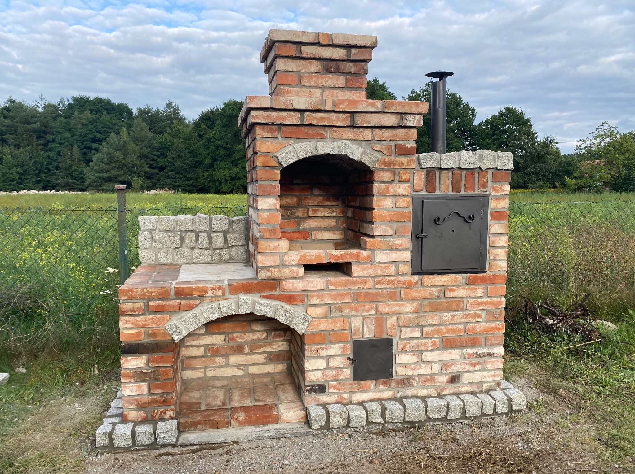 Murowany grill ogrodowy z cegły klinkierowej, z elementami z kamienia naturalnego, na tle zieleni. Widoczny komin i metalowe drzwiczki.
