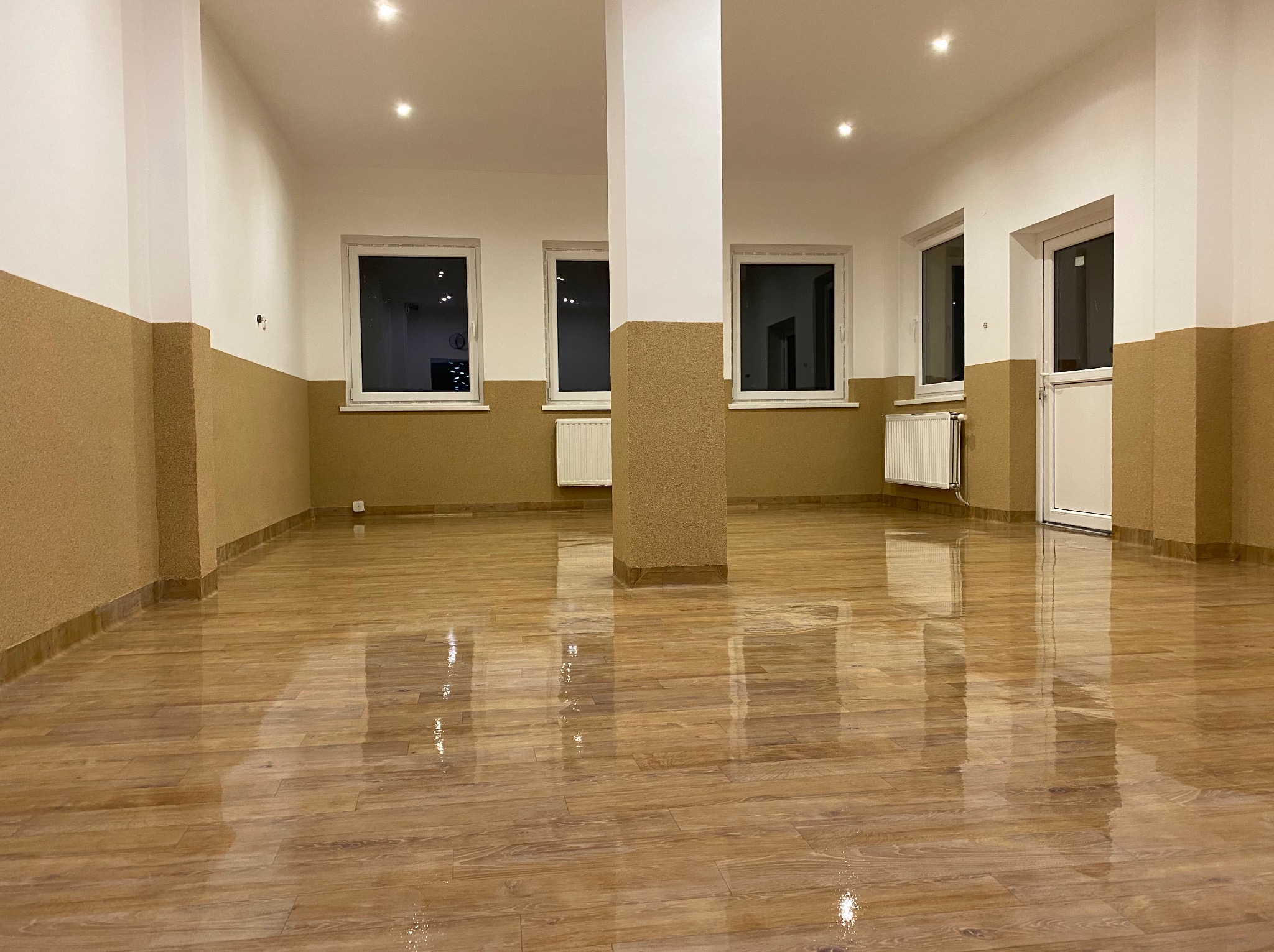 Wykończone wnętrze: błyszcząca drewniana podłoga, ściany z dekoracyjnym wykończeniem do połowy wysokości, białe okna i drzwi. Widoczne grzejniki i kolumna.