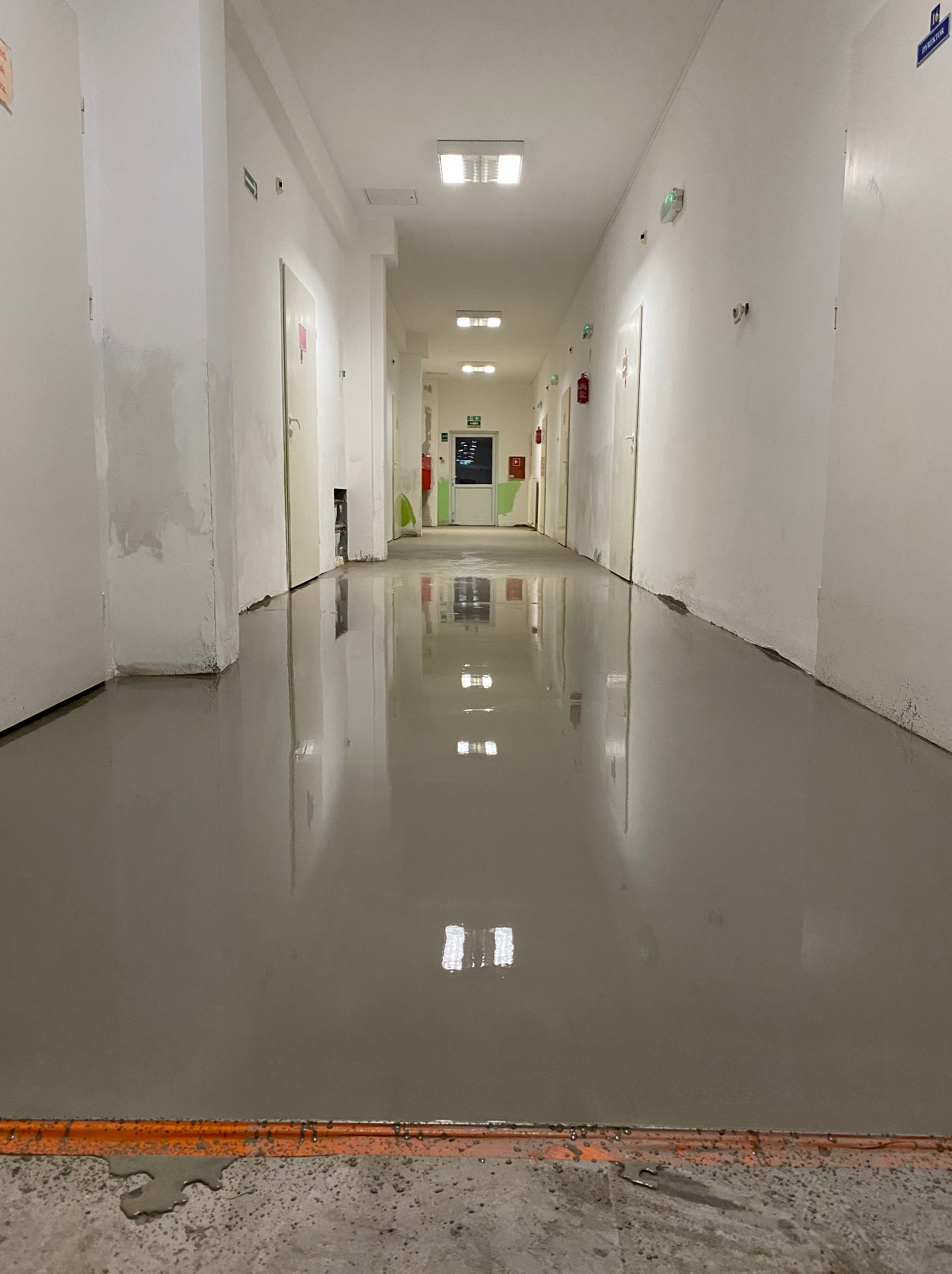 Wypoziomowana, szara posadzka w długim korytarzu z białymi ścianami. Widoczne drzwi i oświetlenie sufitowe. Na podłodze widoczne odbicia światła.