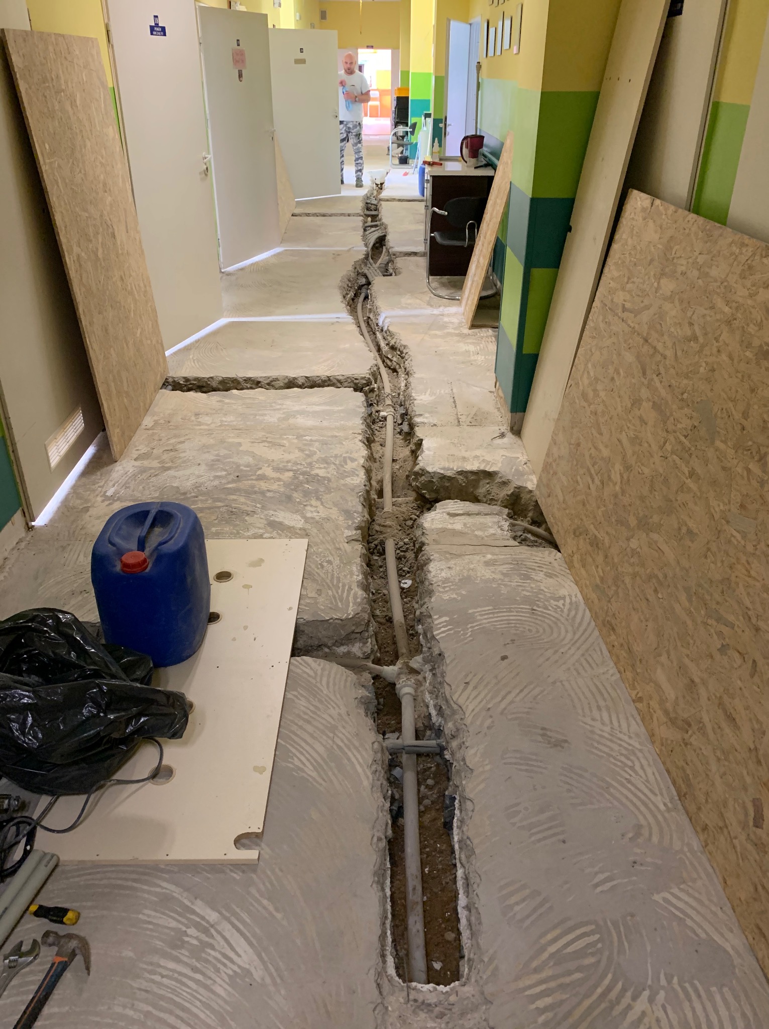 Korytarz w trakcie prac instalacyjnych: wykopane bruzdy w podłodze, widoczne rury, narzędzia i materiały budowlane. W tle osoba wchodząca do pomieszczenia.