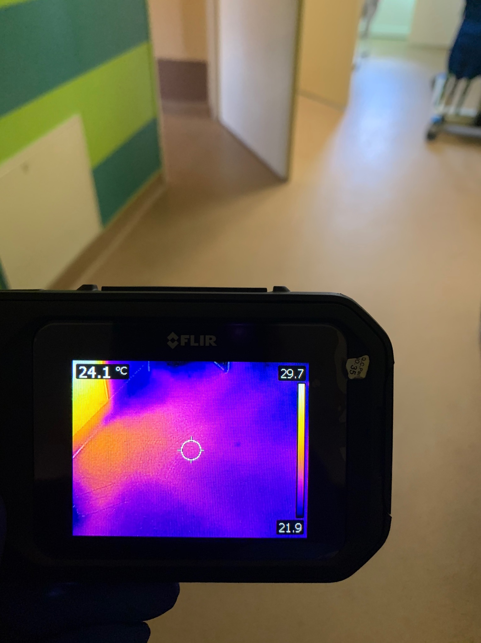 Termowizja podłogi w korytarzu, widok z kamery FLIR. Na ekranie rozkład temperatur oznaczony kolorami, od 21.9 do 29.7 stopni Celsjusza. W tle ściana z zielonymi pasami.