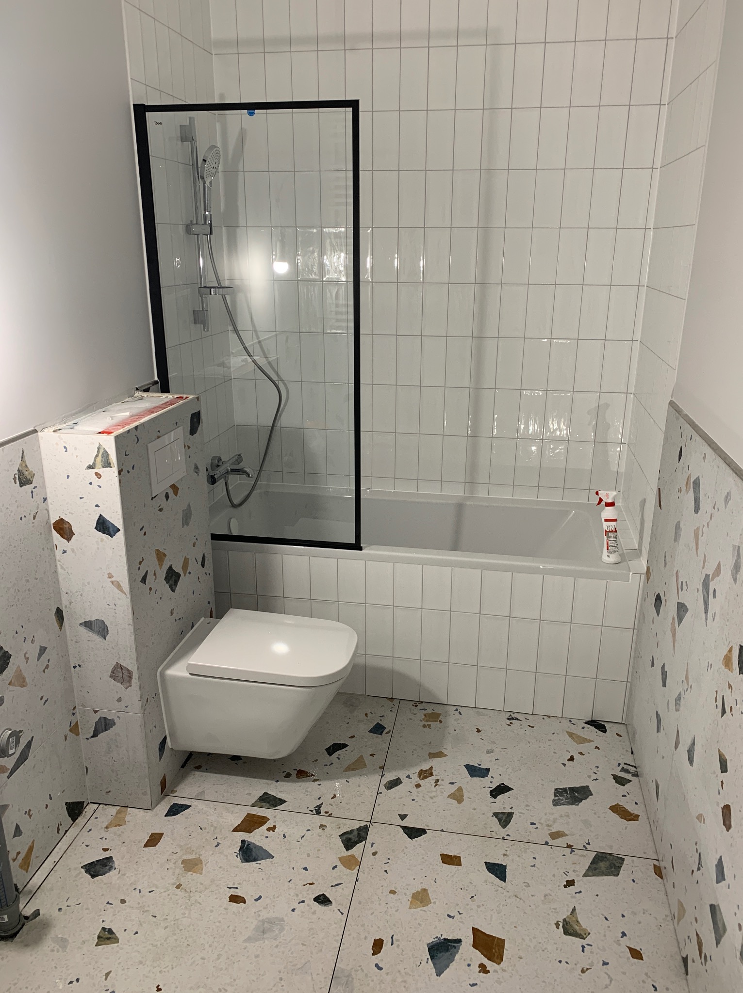 Wykończona łazienka z białą armaturą i płytkami lastryko na podłodze i ścianach. Kabina prysznicowa z czarną ramą i białe, prostokątne płytki na ścianach w strefie kąpieli.