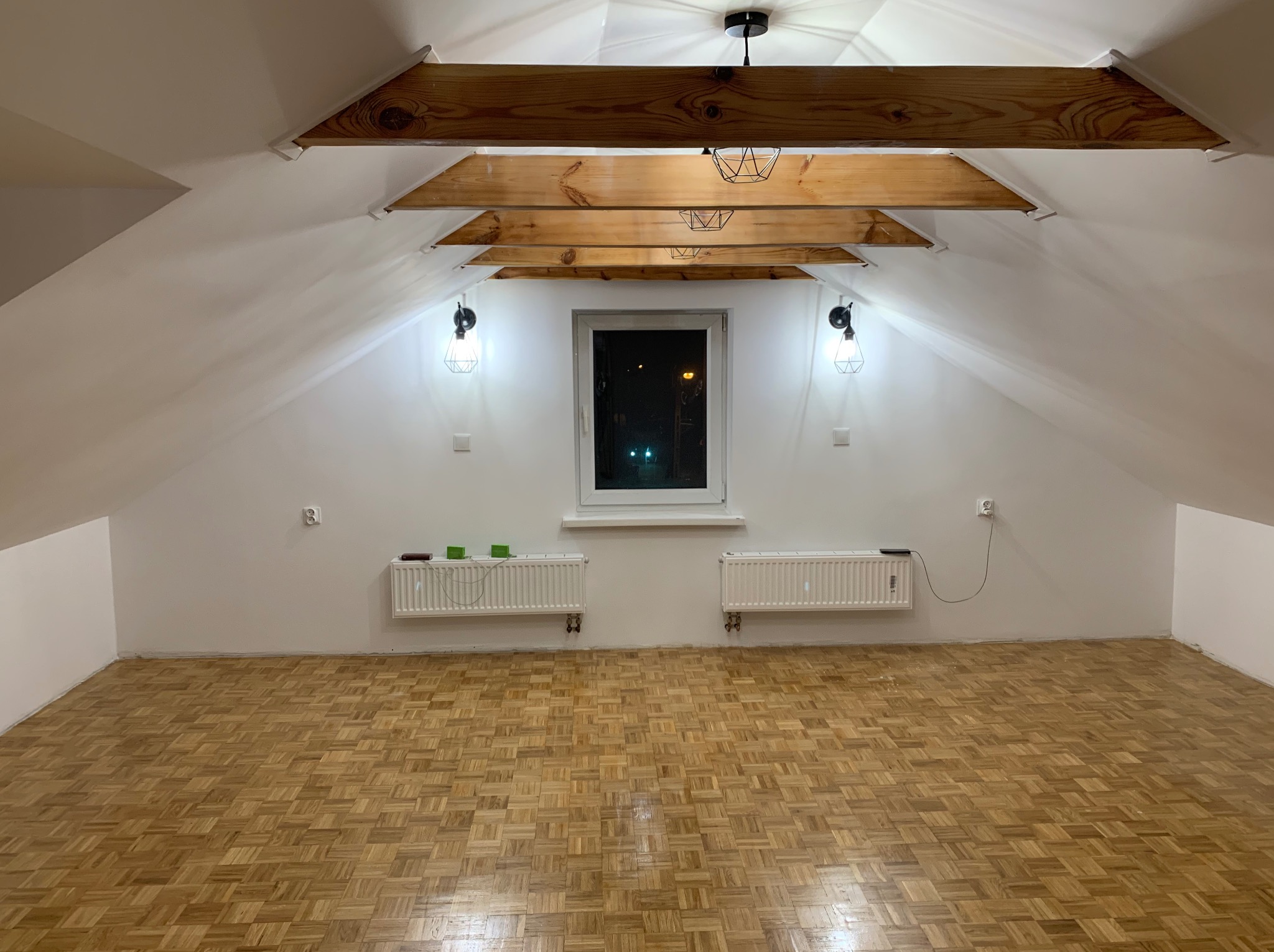 Poddasze po remoncie: parkiet w jodełkę, białe ściany, drewniane belki stropowe z lampami, okno. Widoczne grzejniki i gniazdka elektryczne. Noc za oknem.